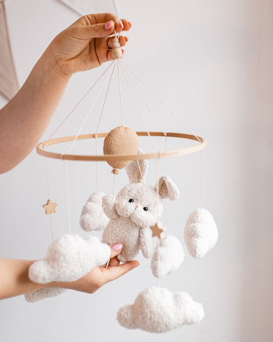Hand holding a baby mobile with a teddy bear, clouds, and stars against a light background